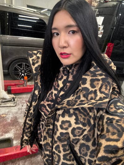 The image depicts a female in an indoor automotive workshop or garage in Herentals, Belgium.

The subject is a female of East Asian descent, positioned centrally and looking directly at the camera. She has long, straight, dark hair with a center part. Her makeup includes prominent red lipstick. She is wearing a padded leopard print jacket over a matching high-collared, zippered garment. The jacket features a dark, stylized emblem on the upper right sleeve.

In the background, to the left of the subject, a dark grey van/minibus with black multi-spoke alloy wheels is visible. Below the van, a red metal vehicle lift arm is present on a concrete floor. An orange and black aerosol can is situated near the lift arm. To the right, the rear section of a second dark vehicle, likely a van or SUV, is partially visible, showing a red taillight. The environment features a utilitarian concrete floor and artificial overhead lighting reflecting on the vehicles.