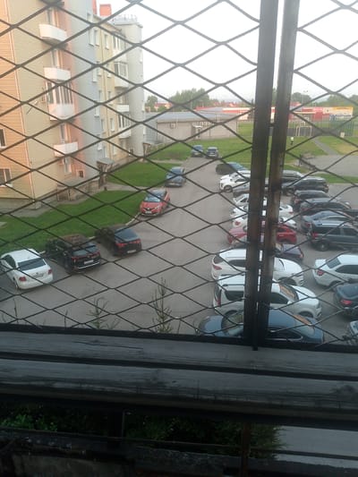 The image shows a view through a wire fence of a residential area in Polysayevo, Russia. In the foreground, a wooden railing is visible. Beyond the fence, a multi-story apartment building with beige and light blue siding, white balconies, and numerous windows is situated to the left. A parking lot with several cars is in front of the building. Visible vehicles include a white Volkswagen Polo, two dark-colored sedans, and a red Renault Duster. Further in the background, more cars are parked in rows, and commercial buildings with red and white facades are present. The sky is overcast. The ground is paved with asphalt in the parking areas and covered with grass in some sections.