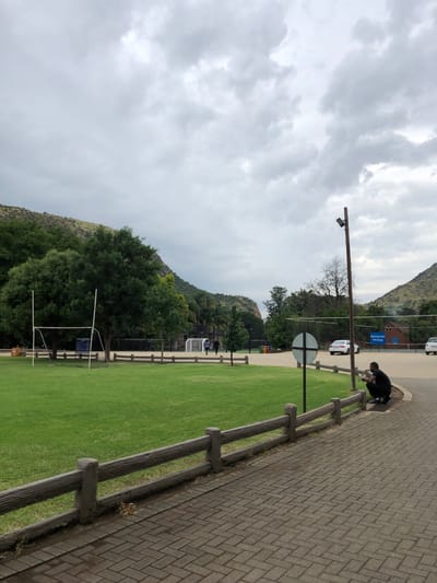 The image captures an outdoor scene under an overcast sky, with the main vantage point looking across a grassy field towards hills in the distance. The image is oriented vertically with the horizon line running vertically down the center.
Starting from the right side of the image (which would be the bottom if rotated correctly), a paved pathway made of interlocking brick pavers stretches into the distance, slightly curving. A low wooden fence with two horizontal rails runs parallel to the pathway, separating it from a large, bright green grassy field.
In the mid-ground, on the paved pathway, a person dressed in dark clothing is crouched down near the fence. Further into the scene, beyond the grassy field, there appears to be a road or parking area with at least two white vehicles visible. A tall, dark pole with a fixture (possibly a light or camera) extends upwards from this area. A circular white sign with a red border, resembling a "No Entry" or similar warning sign, is also visible near the road.
In the background, behind the road and some trees, there's a building with a blue sign on its facade. The text on this blue sign reads "Beginner Cycling". What appears to be a set of white goalposts for a sport like soccer or American football can be seen on the grassy field, closer to the left side of the frame. More people are discernible in the distance on the field.
The far background is dominated by large, undulating hills or mountains covered with a mix of green foliage and drier, lighter-colored vegetation. The sky above is completely filled with thick, grey clouds, indicating an overcast or cloudy day, and casting a soft, diffused light over the landscape. The overall setting suggests a recreational or sports complex situated within a natural, hilly environment.