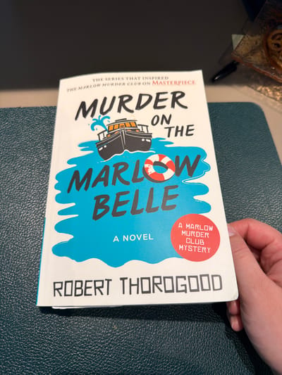 The image shows a close-up, angled view of the book cover for "Murder on the Marlow Belle" by Robert Thorogood. The cover features bold, white text against a bright blue background that mimics water. The title "MURDER ON THE MARLOW BELLE" is prominently displayed. A graphic of a ship with a lifebuoy is also visible, adding to the nautical theme. A small red circle on the upper right side of the cover contains text identifying it as "A MARLOW MURDER CLUB MYSTERY." On the left side of the cover, smaller text reads, "THE SERIES THAT INSPIRED THE MARLOW MURDER CLUB ON MASTERPIECE."

The book is held by a person's hand, with fingers visible at the top of the frame, suggesting the book is being presented or examined. The background consists of a textured, dark surface, possibly a table or desk, with a hint of another object in the upper left corner that appears to be food in packaging. The lighting suggests an indoor setting. The overall presentation is clean and focused on the book cover.