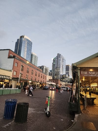 The image presents an urban street scene at dusk within Pike Place Market, Seattle, United States. In the foreground, two cylindrical trash receptacles, one blue and one dark green, are positioned on the left side of a cobblestone street. Adjacent to them, a lime-green electric scooter is parked next to an orange traffic cone. Numerous pedestrians are distributed across the cobblestone street and sidewalks, walking or standing.

On the left, a multi-story building with a red brick facade and visible windows features a "STARBUCKS" sign at its street level. On the right, a market stall or establishment under an awning displays a partial sign reading "THE NOR" and "LOCALLY HANDMADE." This area exhibits interior lighting and a bench with seating. In the midground, a dark sedan and a red car with illuminated brake lights are present on the street, along with a white limousine-like vehicle further in the distance. The background is dominated by several tall, modern skyscrapers with reflective glass exteriors, many showing illuminated interior lights, set against an overcast, light grey sky. A small, dark object, consistent with an aircraft, is faintly visible in the upper left portion of the sky.