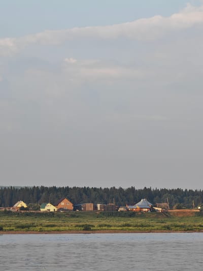The image depicts a rural scene with a wide river in the foreground, reflecting the soft light of the sky. In the middle ground, a densely forested area forms a dark green horizon line. Along the crest of the bank, overlooking the river and forest, is a small settlement of houses. These structures appear to be typical rural dwellings, some with visible chimneys and varied rooflines. The architecture suggests a temperate climate. The overall atmosphere is serene and suggests a peaceful, remote location. The location is identified as the city of Lesosibirsk, Russia, a city known for its timber industry and location in Siberia, which aligns with the natural landscape depicted.