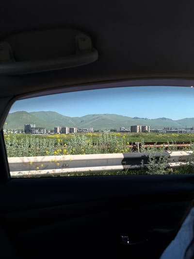 The image, taken from inside a vehicle, shows a view through the car window of a city with a backdrop of rolling green hills under a clear blue sky. In the foreground, a metal guardrail, partially rusted and painted with white and black stripes, separates the road from a field of green grass dotted with small yellow flowers. Beyond the guardrail, a residential area is visible with several mid-rise apartment buildings. One prominent building in the mid-ground appears to be under construction, with scaffolding and a crane. The city of Sevan, Armenia, is known for its beautiful natural setting, with the Sevan Lake and surrounding mountains, and this scene aligns with the typical landscape of the region, suggesting the image was captured from a vantage point overlooking the city and its natural environment.
