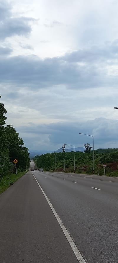 The image depicts a road in Khian Sa, Thailand. The road is asphalt with a white dividing line. The sky is cloudy. Trees and vegetation line the road. There are streetlights along the road. A yellow sign is visible on the side of the road, as well as another traffic warning sign farther down the road. In the distance, there are two cars traveling on the road.