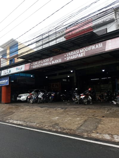 The image depicts a street-level view of an automotive service and repair shop located in Cipendawa, Indonesia, under an overcast sky. The shop occupies the ground floor of a multi-story commercial building, with upper floors featuring windows and blue louvered coverings. Numerous black overhead power lines are visible crisscrossing above the building.
In the foreground, an asphalt road with a white dividing line is visible. Directly in front of the shop, a concrete apron or sidewalk, showing signs of wear and dirt, extends from the road. The shop itself has an open front with a dark interior, illuminated by several ceiling lights. A long banner stretches across the facade, displaying services in Indonesian: "TUNE UP & BORE UP," "SERVICE KARBU & INJEKSI" (Carburetor & Injection Service), "VARIASI MODIFIKASI" (Modification Variations), and "SPAREPART" (Spare Parts). A "MICHELIN" logo is visible on an orange corrugated metal shutter partially open on the far left of the shop. Faint "RKN" lettering is visible on an interior wall.
Inside and immediately in front of the shop, a white Honda crossover vehicle is parked on the left. Approximately 15 to 20 motorcycles and scooters of various models and colors (predominantly black) are parked in rows, filling much of the shop's front area and extending onto the concrete apron. In the rightmost bay of the shop, at least two individuals are present. One person is crouched or kneeling on the ground, actively working on a motorcycle, while another individual is vaguely visible nearby.