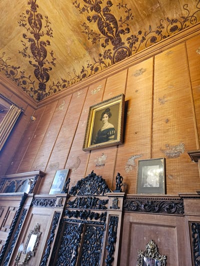 The image shows a corner of an ornately decorated interior, likely a room within a historic building. The location is potentially in Алупкинский городской совет, Ukraine. The corner is defined by walls and ceiling panels covered in wood.
The wall on the left is adorned with dark brown floral patterns on a light brown surface. A portrait in a gold frame hangs on the adjacent wall, which is constructed from narrow wooden planks. These planks also feature faint, lighter-colored floral patterns. Another framed picture is above and to the right of the portrait.
A complexly carved wooden structure is on the right, showing darker wood and intricate designs. Additional framed pictures are visible on this structure. The overall impression is of an elaborate, historical interior.