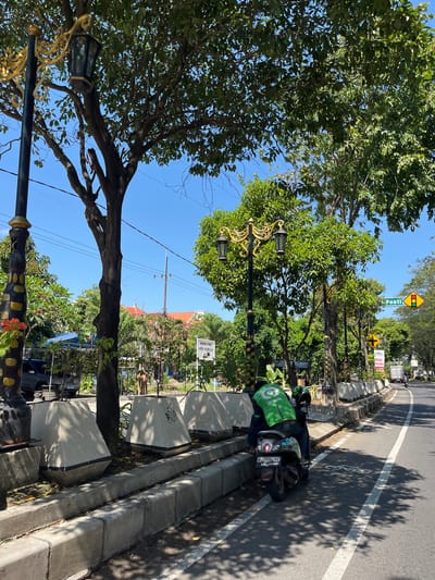 The image depicts a street scene in Pagerwojo, Indonesia, on a clear day. In the foreground, a person wearing a green "Gojek" uniform rides a scooter along a road. The scooter has a license plate that reads "W 2606 VC". The scooter is positioned on a paved road with white lane markings. Adjacent to the road is a concrete barrier and a row of planter boxes.
Several street lamps with ornate gold accents are visible, some partially obscured by trees. The trees are lush and green, with dense foliage. In the background, there are buildings with terracotta roofs and more greenery. Traffic signs are also present, including a diamond-shaped yellow sign with a black arrow and a green sign that says "Pontianak". The overall environment suggests a well-maintained urban or suburban area. The lighting indicates it is daytime, with strong shadows cast by the trees.