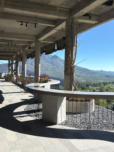 The image depicts an outdoor structure with a view of mountains in Kedisan, Indonesia. The structure features a concrete frame with round concrete posts, some of which support canopies and plant boxes containing flowers. There are small lights mounted on the concrete walls. Alongside the structure is a walkway covered with gray stones and patterned gray tile, casting shadows. A railing separates the walkway from a section covered in smaller gray pebbles. The view beyond the structure shows a mountainous landscape with vegetation under a clear blue sky.