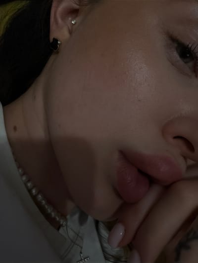 A close-up, angled shot depicts a woman's face, positioned with her nose and mouth centered. Her right hand is partially obscuring her mouth. Her eye is visible on the upper left side of the frame. She has light skin and her lips are slightly parted. She is wearing a pearl necklace. Her ear is visible on the lower left side of the frame, adorned with a black heart-shaped earring and a small stud earring above. A tattoo is partly visible on the upper right. The location is Воскресенские Усадьбы Кп, Russia.