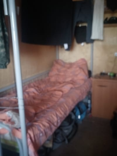 The image depicts a dimly lit, cramped living space, likely a dormitory room or a small dwelling in Vybenka, Russia. The focal point is a narrow bed with a salmon-colored comforter, appearing somewhat disheveled. To the left of the bed, a white metal frame of what appears to be a bunk bed is visible. Above the bed, dark clothing, possibly a sweatshirt and pants, is hung on a rack. A pale grey garment is also hanging nearby.

On the right side of the image, a wooden cabinet with a drawer is situated. On top of the cabinet, a small green object, possibly a bottle or container, is visible. The walls of the room are a light brown, wooden paneling. Various bags and dark objects are scattered on the floor beneath and to the side of the bed, suggesting a lived-in, possibly untidy environment. The overall impression is of a functional, but spartan, living quarters.