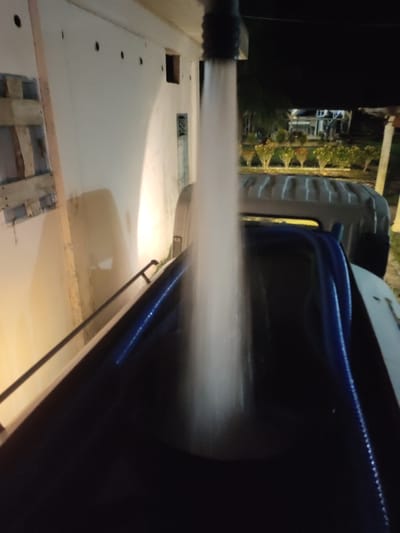 The image shows a stream of water falling from a dark nozzle into a dark blue container. The container appears to be the bed of a truck. In the background, there is a building with white walls and some windows. There are also some plants and trees visible in the distance. The scene is illuminated by artificial lights. The overall impression is that the water is being pumped into the truck bed. It is nighttime and the location is Beuracanrata, Indonesia.