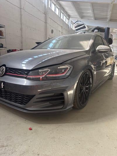A dusty, dark gray Volkswagen Golf GTI is parked indoors, likely in a garage or workshop in Schkopau, Germany. The car is angled, with the front left side most visible. It has a honeycomb grille with a red stripe, red accents on the headlights, and black multi-spoke wheels. The car's interior is visible through the dusty windows, and its windshield reflects the skylights in the ceiling. Another car, a white SUV, is partially visible in the background, elevated on a lift. The environment appears to be an industrial or automotive repair space, with concrete floors, white walls, and exposed ceiling beams and pipes. There are shelves with various items on the left side of the image. A small red object, possibly a bottle cap, is on the floor near the front of the car. There are no people visible in the image.