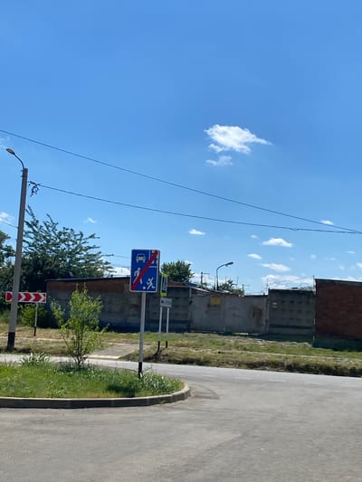 The image shows a street scene with a blue traffic sign indicating that motor vehicles and pedestrian crossings are prohibited. The sign is mounted on a metal pole. To the left of this sign, another pole supports a red and white directional arrow sign. A small green tree grows in a grassy area in the foreground, next to the road. In the background, a brick wall and a grey concrete fence are visible. Overhead power lines are strung across the bright blue sky, which has a few white clouds. The setting appears to be an urban or suburban area. The image was taken in the city of Maykop, Russia.