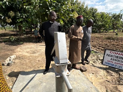 In Afogo, Nigeria, three men stand near a water pump. The man on the left, wearing glasses and a black robe with a white collar, stands closest to the pump. To his right, a man in a brown robe and a red hat is holding a document. The third man, in a grey robe, stands slightly behind and to the right of the man in brown. The water pump is a metallic structure mounted on a concrete base. In the background, there are trees and tilled earth. A sign with red and blue lettering is visible on the right side of the image. The ground is dry and dusty.