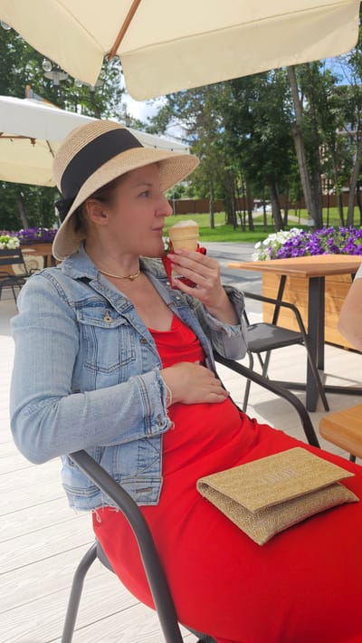 A woman with blonde hair, wearing a straw hat with a black ribbon, a denim jacket, and a red dress, is sitting on a chair outdoors and eating an ice cream cone. She is holding the cone to her mouth with her right hand and resting her left hand on her stomach. A woven clutch bag is placed on her lap. In the background, there are trees, a wooden table, and chairs, suggesting an outdoor seating area, possibly at a cafe or restaurant in Alikonovka, Russia. The ground is made of white wooden planks, and there are outdoor umbrellas providing shade.