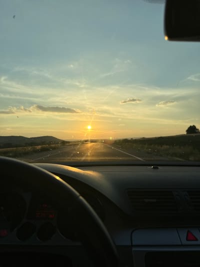 The image shows a view from inside a car looking out at a sunset over a highway. The sun is low on the horizon, casting a warm glow across the sky and the road. The road stretches ahead, with rolling hills on either side. The car's dashboard and steering wheel are visible in the foreground. There are some dust or water spots on the windshield. The image does not provide any information that would allow for identification of the location in Vladimir, Bulgaria.