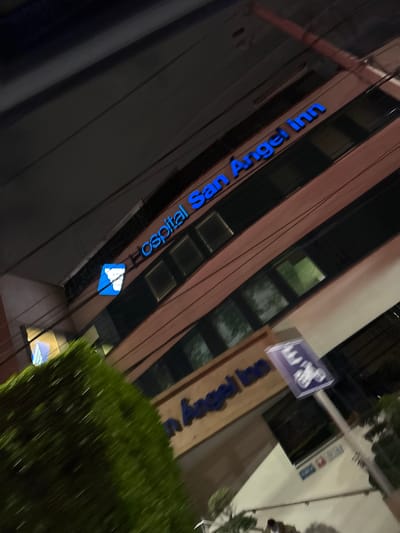 The image shows the exterior of the Hospital San Angel Inn, located in Tizapan, Mexico. The building is multi-story, with the name "Hospital San Angel Inn" displayed vertically in blue lettering on the facade. Some blue and white symbols are visible below the hospital's name on the building. A sign with blue figures is mounted on the building near the top. Green bushes are near the building's sign. It appears to be nighttime.