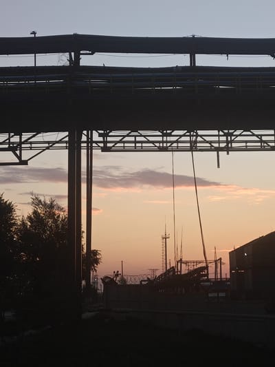 The image depicts an industrial landscape at dusk or dawn, with a clear sky transitioning from light blue to a soft orange and pink hue. In the foreground, silhouetted against the sky, are multiple layers of industrial infrastructure, including pipes and structural supports, creating a dominant horizontal and vertical pattern. A large tree with dense foliage is visible in the left mid-ground, also in silhouette.
Further into the mid-ground, various industrial elements are present, including what appears to be a power pylon or communication tower, barbed wire fencing, and a building with a dark roofline on the right. Several wires or cables are suspended from the overhead structures, adding to the industrial feel. The overall scene suggests an operational or decommissioned industrial site. The location is identified as Дүңгілағаш, Kazakhstan. There are no people visible in the image.