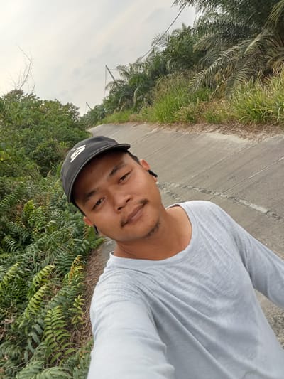 A man wearing a black baseball cap and a light gray long-sleeved shirt is taking a selfie outdoors. He has a short mustache and goatee and is wearing black wireless earbuds. The man is standing on a paved road that curves to the right and is bordered by lush green vegetation on both sides. In the background, palm trees are visible, and the sky is overcast. The road appears to be a rural path, possibly in an agricultural area.