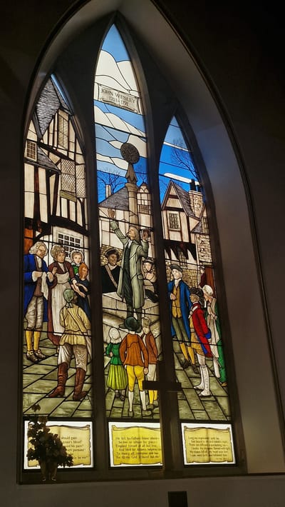 This is a photograph of a stained-glass window depicting John Wesley, the founder of Methodism. The window is located in the Llandudno United Methodist Church in Llandudno, United Kingdom. The window is divided into multiple panels. The top panel features the inscription "JOHN WESLEY 1703-1791". The central panel shows Wesley preaching to a crowd in what appears to be a 17th or 18th-century town square. He is depicted standing on a raised platform, holding a book and raising his arm, as if addressing the onlookers. The surrounding buildings are in a Tudor architectural style. The lower panels contain text which appears to be quotes from Wesley's hymns. The window is set within a gothic archway. To the lower left of the window, there is a small vase with artificial flowers.