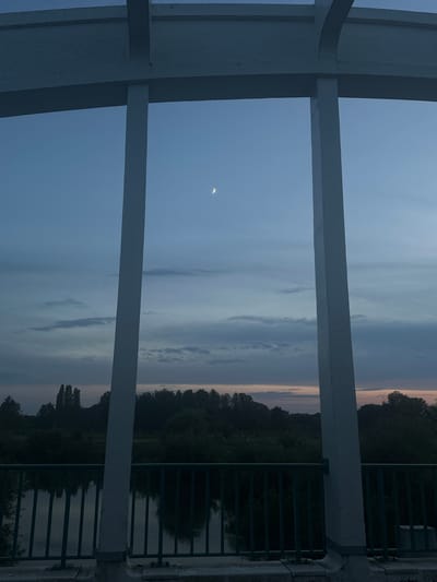 The image depicts a dusky sky over a body of water and a treeline, as viewed through the structural elements of a bridge. A sliver of the crescent moon is visible in the upper portion of the frame. The foreground features the white concrete structure of the bridge and a dark green metal railing. Beyond the railing, the water reflects the muted colors of the twilight sky. A dense line of trees forms the horizon, with a few taller trees interspersed. The overall scene suggests a peaceful, natural setting observed from an elevated vantage point within the city of Sempigny, France, likely during the early evening hours. There are no people or animals visible in the image.