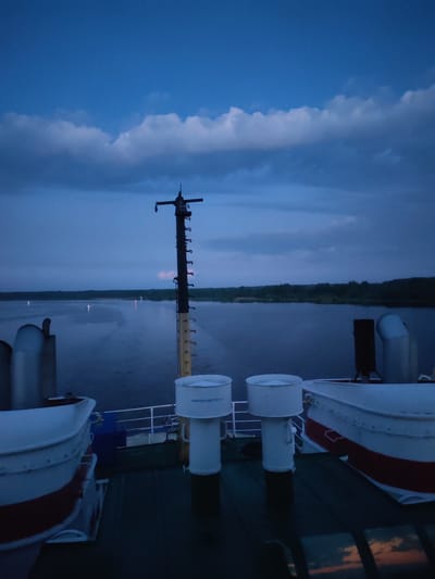 The image shows a view from the deck of a vessel, likely a ship or large boat, on a body of water in what appears to be the city of Irdomatka, Russia. The scene is set during dusk or twilight, as evidenced by the dim lighting and the color of the sky. The sky is predominantly blue, with clouds visible. The water is calm and reflects the ambient light. On the deck of the vessel, there are lifeboats and cylindrical structures, possibly exhaust vents or navigational equipment. A tall mast with ladder rungs and antenna is positioned in the center of the view. On the far bank of the body of water, there is a line of trees forming a treeline, suggesting a forest or park area. Several lights can be seen on the water, presumably from other vessels or structures along the shore.
