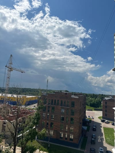 This image depicts an outdoor scene in Rubtsovo, Russia, likely taken during the daytime under a partly cloudy sky. The perspective is from a high vantage point, looking down and across an urban landscape.
The main subjects are several multi-story brick buildings, some appearing to be residential or commercial. One prominent building in the foreground has a red brick facade with numerous rectangular windows. Adjacent to it, another building is partially visible with yellow and blue siding.
In the middle ground, a construction site is evident, featuring two tall cranes, one of which is a lattice boom crane. Cars are parked along paved pathways and green grassy areas surround some of the buildings. Trees with green foliage are scattered throughout the scene, with some bare branches from a tree in the immediate foreground.
The sky is a mix of bright blue and large, fluffy white clouds, suggesting fair weather. The overall impression is one of ongoing development or construction in a moderately built-up area. No people are clearly visible. There is no discernible text in the image.