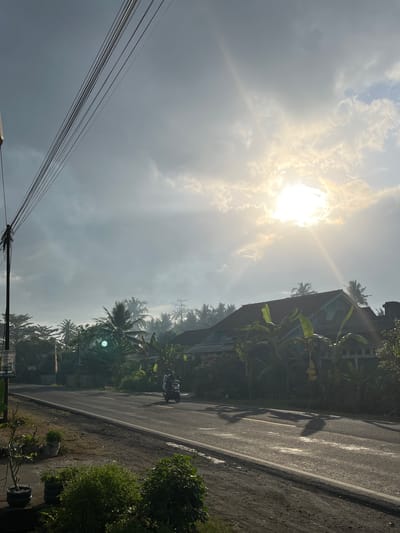 In Tambakreja, Indonesia, a street scene is captured during what appears to be either sunrise or sunset, with the sun's bright rays piercing through a cloudy sky. Overhead, a bundle of electrical wires runs diagonally across the frame, attached to a utility pole on the left side of the image. The road, paved with asphalt and marked with a yellow centerline, stretches into the distance. A motorcycle with two occupants is traveling along the road. On the right side of the road, a building with a reddish-brown roof and a pale blue facade is visible, surrounded by lush green vegetation including banana trees and palm trees. The foreground features a dirt shoulder with potted plants. The overall atmosphere is serene and rural, with natural light dominating the scene.
