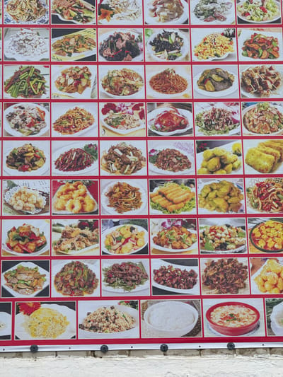 The image displays a vibrant, large-format menu board showcasing a variety of Chinese dishes. The board is divided into a grid of numerous individual photographs, each featuring a different culinary preparation. These photographs are arranged in rows and columns and are bordered by red frames. The dishes themselves appear to be well-lit and appetizing, with a diverse range of ingredients and cooking styles evident. Many of the dishes feature stir-fried vegetables, meats, and noodles, while others include fried items, rice dishes, and soups. The backdrop of the menu board is white, with a prominent red border. Below the menu board, a textured white brick wall is visible, with two black circular fasteners attached to the wall at the bottom of the menu board. The scene is brightly lit, suggesting an outdoor setting or a well-lit indoor space. The overall impression is that of a restaurant menu designed to attract customers with appealing visual representations of their offerings. The location is specified as Chuguyevka, Russia.
