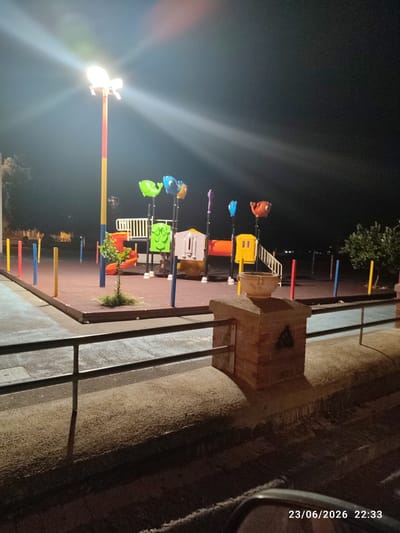This media file depicts a brightly lit playground at night, likely in Scordia, Italy, as indicated by the timestamp "23/06/2026 22:33". The scene is illuminated by a tall, multi-colored pole light that casts strong beams and lens flares across the dark sky. The playground equipment is colorful, featuring what appear to be stylized animal-shaped swings or decorations in shades of green, blue, and orange, along with slides and climbing structures. The ground of the playground is covered with red rubber matting.
In the foreground, a low brick structure with a circular planter on top is visible, along with a metal railing. The background shows more of the playground with colorful poles marking its perimeter, and a glimpse of trees and distant lights, suggesting an urban or suburban setting. The overall atmosphere is one of a quiet, well-maintained public space after hours. There is no activity or people visible in the frame, giving the scene a sense of stillness. The image is taken at night, with the artificial light being the primary source of illumination. The weather appears clear, as indicated by the sharpness of the light beams.