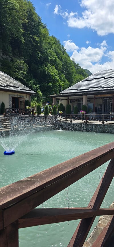 The image depicts a serene outdoor scene in Babugent, Russia, likely during the daytime under a clear blue sky with scattered white clouds. The setting is a waterside establishment, possibly a restaurant or resort, nestled against a lush, tree-covered mountainside.

In the foreground, a weathered wooden railing forms a diagonal line across the frame, suggesting a viewing platform or walkway overlooking a body of tranquil, greenish water. A water fountain with a blue nozzle is actively spraying water, creating a dynamic, misty effect.

Along the edge of the water, there's a building with a dark, tiled roof and a stone facade. A walkway or patio area, bordered by a metal railing and decorative potted plants, runs alongside the building. A few people are visible on this walkway, appearing relaxed as they observe the surroundings. One person, wearing a blue cap, stands near a small tree.

The mountainside behind the building is densely covered with vibrant green trees, indicating a natural and possibly remote location. The overall atmosphere is peaceful and inviting, capturing a moment of calm amidst a beautiful natural landscape. There is no visible text in the image.