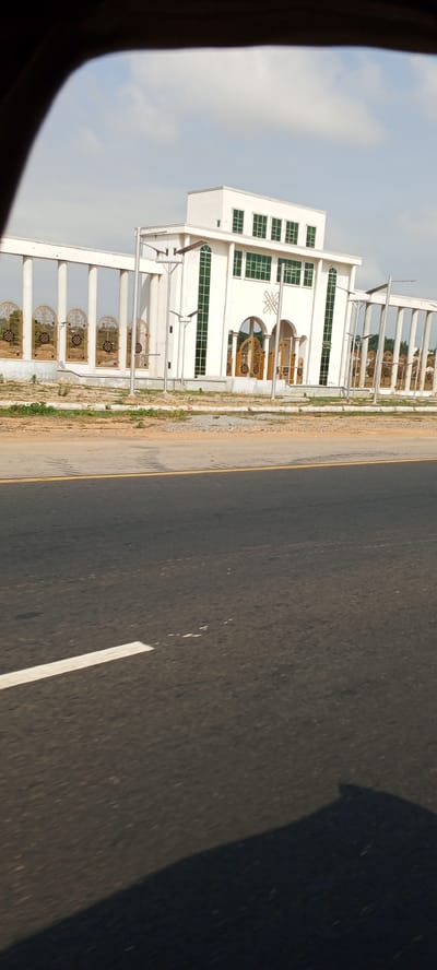 A low-angle shot captures a white building with green-tinted windows and double doors, framed by a colonnade. The building stands adjacent to a paved road marked with a white line. Streetlights are present in front of the building. The location is presumed to be Uke, Nigeria. No people or interactions are visible.