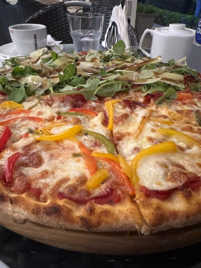 The image shows a close-up of a pizza with a variety of toppings, including melted cheese, sliced bell peppers in red, yellow, and green, and possibly some meat. A side salad with greens, thinly sliced vegetables, and a drizzle of balsamic glaze is also visible, placed next to the pizza. In the background, there are hints of an outdoor setting with glassware and what appears to be a white ceramic mug with text. The overall scene suggests a meal being served, possibly at a restaurant. The lighting in the image is somewhat dim, indicating it might be evening or nighttime. The presence of glassware and cutlery hints at a dining establishment. The visible text on the mug reads "CANONERO". The context provided indicates the location is Malé, Maldives. No people are visible in the frame, and the focus is entirely on the food and immediate surroundings.