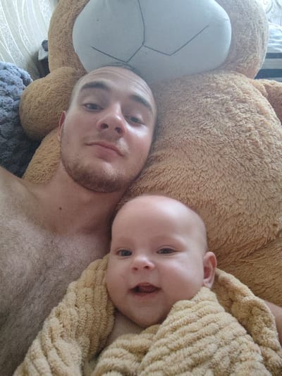In Okhtyrka, Ukraine, a young man and a baby are pictured lying down with a large teddy bear. The man is looking at the camera with a slight smile, while the baby is looking away. The baby is wrapped in a soft, fuzzy blanket. The background is blurry, but it appears to be a room with striped wallpaper. The overall scene is one of warmth and affection.