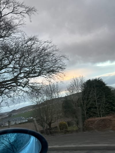 The image depicts a rural scene in Kirkton of Auchterhouse, United Kingdom. The sky is overcast with a hint of sunlight breaking through the clouds. Bare trees dominate the foreground. A road lined with hedges and other foliage runs along the right side of the frame. In the distance, a building can be seen. A car side-view mirror is visible in the bottom right corner, reflecting the trees.