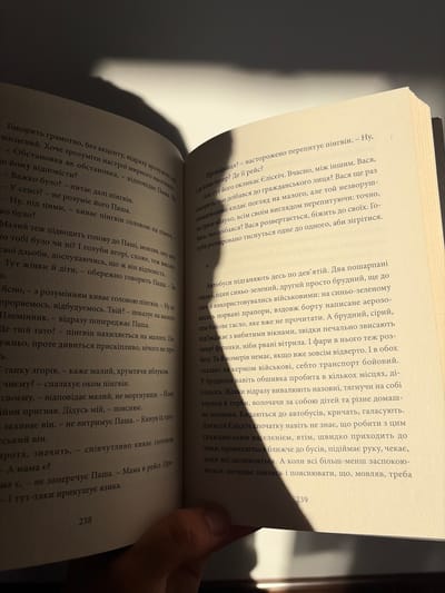 The image shows a close-up view of an open book, illuminated by a harsh overhead light casting a strong shadow across the pages. The book appears to be a novel, with dense text filling both visible pages. The left page contains dialogue and narrative, depicting a scene with characters named Pasha and a character referred to as "pingvin." The right page continues the narrative, describing a group of people, possibly a family, interacting with a bus. The overall scene is indoor, likely a room in a residential building. No specific landmarks or elements are present to indicate a location within Hanniv, Ukraine. The image is shot from a high angle, looking down at the book. The focus is sharp on the text, with a shallow depth of field. The lighting creates a dramatic contrast between the illuminated text and the shadowed areas of the book and the background. The bottom right corner of the image reveals a human hand holding the book open.