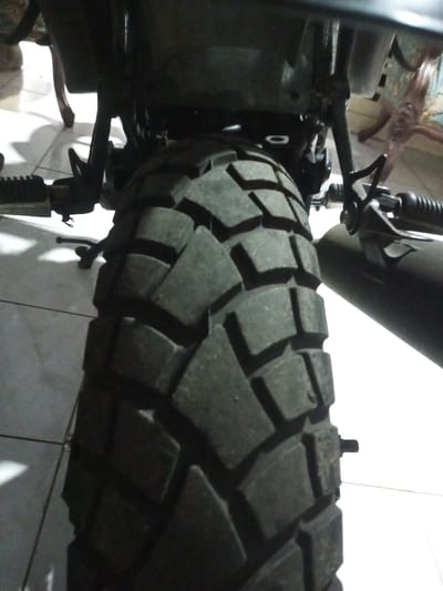 The close-up image showcases a motorcycle's rear tire, presenting a detailed view of its tread pattern. The tire's surface exhibits deep, block-like treads, suggesting an off-road or dual-sport design. The tire is positioned under the motorcycle's frame, with portions of the exhaust pipe and foot pegs visible on either side. The background reveals glimpses of a tiled floor. The motorcycle is stationary, and the focus is clearly on the tire's rugged construction.