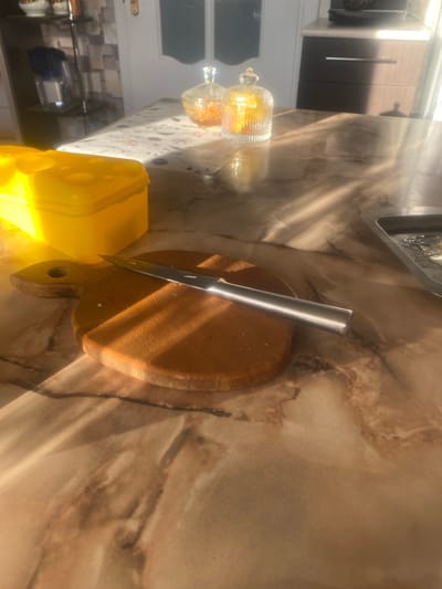 The image shows a close-up view of a kitchen table with a wooden cutting board and a knife. A bright yellow container, possibly a cheese keeper, is positioned to the left of the cutting board. The table has a marble-like pattern and is illuminated by strong sunlight casting shadows. In the background, a white patterned tablecloth is visible, along with two glass jars containing what appears to be orange-colored food items. Further in the background, shelves with kitchen items and a cabinet can be seen. The overall scene suggests a domestic setting, likely a home kitchen, and the lighting indicates it is daytime. There is no information within the image or accompanying text that would allow for specific identification of the location within the city of Zhodzina, Belarus.