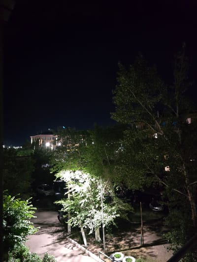 This media file shows a night scene in Shahtinsk, Kazakhstan, likely in a residential area. The image is oriented vertically, with the top of the frame appearing on the left. In the foreground, the branches and leaves of trees are prominent, illuminated by artificial light sources, creating a dappled effect on the surrounding area.
Further into the scene, a multi-story building with lit windows is visible, suggesting it is nighttime. Cars are parked on a paved surface, also illuminated by the artificial lights. The overall atmosphere is dark, characteristic of nighttime. There are no people visible in the frame. The lighting suggests that the image was taken at night, under artificial lights. The scene appears to be an outdoor area adjacent to a building, possibly a courtyard or parking area. No weather conditions are discernible from the image. No text is visible.