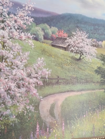 This is a painting of a pastoral landscape, likely a depiction of a rural area in the city of Lyepyel, Belarus. The foreground features blooming pink trees, their branches laden with delicate blossoms. A dirt path winds its way through a grassy hillside dotted with small yellow flowers. In the midground, a rustic wooden house with a dark roof sits on the crest of the hill, surrounded by more blooming trees, some with white flowers and others with vibrant red foliage. Further back, rolling green hills rise towards a hazy blue sky with soft white clouds. The overall impression is one of peaceful natural beauty.