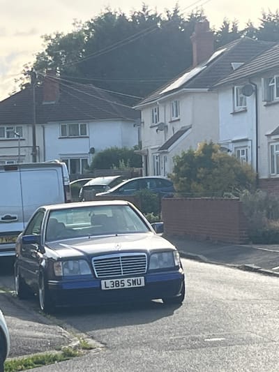 The image depicts a street scene in Maidenhead, United Kingdom. A dark blue Mercedes-Benz with the license plate "L385 SNU" is parked on the right side of the road. Behind the Mercedes is a white van with a partially visible license plate ending in "VHG". Several other vehicles are parked further down the street. On the left side of the street are residential buildings. The road appears to be paved and shows signs of wear. Overhead power lines are visible. The image is taken during daylight hours, with overcast skies.