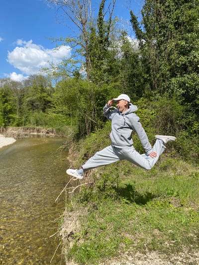 An outdoor, full-length portrait captures a young person striking a dynamic pose beside a clear, shallow river under a bright blue sky. The individual, dressed in a casual grey sweatsuit (hooded sweatshirt and sweatpants), a white baseball cap, grey socks, and white clogs, is shown with one leg bent and lifted, and arms also bent. They appear to be smiling or making a lighthearted expression.
The setting is a vibrant natural landscape in Beregovoe, Russia. Lush green trees and bushes form a dense backdrop, with bright green grass covering the ground where the person stands. The river's clear water reveals a stony bed, flowing gently in the foreground and mid-ground. Above, a vivid blue sky is dotted with fluffy white clouds, indicating sunny and pleasant weather, likely during daytime in spring or summer given the abundant greenery. No text is visible in the image.