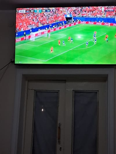 In Kahramon, Uzbekistan, a television displays a live broadcast of a soccer match between the Netherlands (NED) and Sweden (SWE). The scoreboard indicates the Netherlands leading Sweden 4-0 at 56:47 into the game. The screen shows a green soccer field with players in orange and blue uniforms. Spectators wearing orange are visible in the stands, and Coca-Cola advertisements line the field. The television is mounted on a wall above a doorway with two doors. The doors are made of light-colored wood and have dark-tinted glass panes and metal handles. The surrounding wall is light-colored.