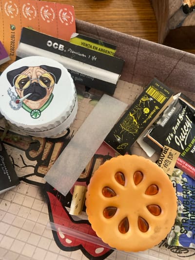 A variety of smoking accessories are arranged on a surface. Items include packs of Juicy Jay's and OCB rolling papers, a grinder with a cartoon dog design, a pack of 3Rayos rolling papers, a plastic toy resembling a pie, and a rolling filter tip. Some of the rolling papers are premium 1 1/4 size. One pack features a strawberry shortcake flavor.