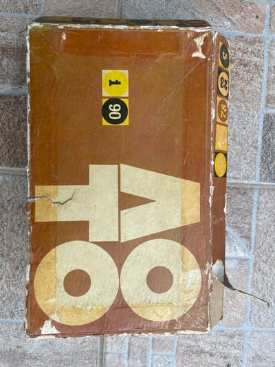 The image depicts a brown, aged cardboard box with the word "ATTO" printed on it in large, stylized white letters. The box appears to be worn, with frayed edges and areas where the surface is peeling or torn. In the top left corner, there are several small, round stickers with numbers printed on them: "5," "23," and "84," and another partly visible. On the left side, there is a yellow sticker with the number "1" and a black sticker with the number "90." A piece of the cardboard is peeling upwards on the upper right corner of the box. The box is placed on a tiled surface with a gray and brown patterned design. The scene is likely set in Byal Izvor, Bulgaria, although this is not explicitly evident from the image alone. There are no people visible in the image.