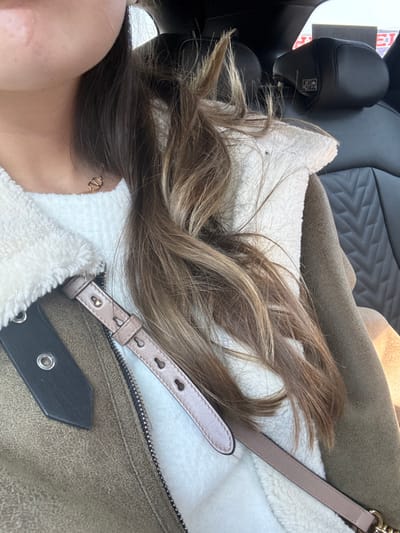 A close-up shot features a woman reclining in a car seat, with her head resting against a white, fuzzy surface. Her long, wavy brown hair cascades across a tan jacket with a fuzzy white lining, and a light pink leather strap lays on top of her jacket. A black leather strap lies on top of the jacket. A portion of her neck is visible, adorned with a delicate gold necklace. The car seat is upholstered in black, with a quilted diamond pattern.