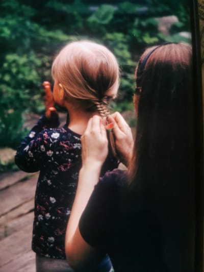 In a dimly lit outdoor setting, two individuals are visible, with their backs mostly to the camera. On the left, a child with light blonde hair is having their hair braided. They are wearing a dark blue long-sleeved shirt with a floral pattern. Their right hand is raised and appears to be holding something, possibly a hair accessory. On the right, an older individual, likely an adult, is braiding the child's hair. They have dark brown hair and are wearing a dark top. A dark blue headband is visible on their head. The background consists of blurred green foliage and what appears to be a wooden pathway or stairs. The overall scene is intimate and suggests a moment of care and interaction between the two individuals. The location in Serov, Russia is not discernible from the image itself.