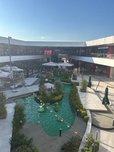 The image shows an outdoor shopping center with a central water feature. In the foreground, a bright green artificial pond has decorative sculptures of a duck and a frog. String lights with exposed bulbs are suspended over the water. Surrounding the pond are meticulously landscaped bushes and trees.

In the mid-ground, there are several outdoor seating areas with tables and umbrellas, suggesting cafes or restaurants. People are seated at some of these tables. Raised walkways and platforms are integrated into the design, connecting different areas of the complex.

In the background, multi-story buildings house various retail stores, identified by signage such as "SPORTIVE" and "SKECHERS." People can be seen walking on the upper levels and inside the stores. The architecture of the shopping center is modern, with white facades and overhead coverings.

The scene is illuminated by natural daylight, indicating it is daytime. The sky is clear and blue. This shopping center is located in Çanakkale Merkez, Turkey.