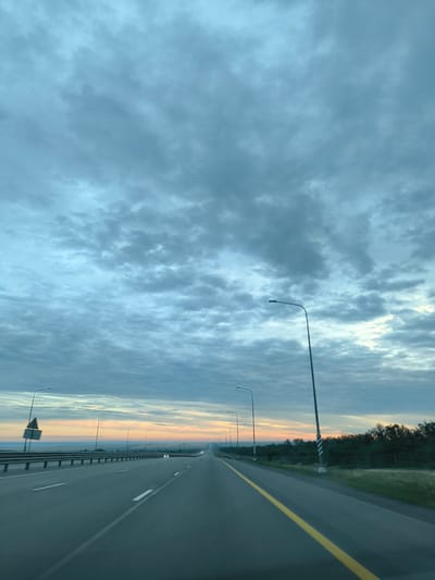A low-angle, vertical shot captures a highway in Staraya Stanitsa, Russia, under a dramatic, cloudy sky. The road stretches into the distance, with a yellow line on the right and white lane markings. Streetlights line the highway, with one prominent lamp post in the foreground on the right. The sky is filled with a dense layer of textured, grayish-blue clouds, with hints of pale orange and pink peeking through at the horizon, suggesting either sunrise or sunset. The landscape on either side of the highway consists of a grassy verge and trees on the right, and a metal guardrail with a warning sign on a pole on the left. No people or vehicles are clearly visible.