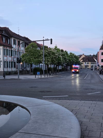The image captures a street scene in Ansbach, Germany, near the Martin-Luther-Platz. In the foreground, a curved, concrete fountain edge is visible, with water reflecting the sky and surrounding structures. The street itself appears wet, possibly from recent rain, with lane markings and a manhole cover visible. On the left side of the street, traditional German buildings with red-tiled roofs, white facades, and green shutters line the pavement. Trees with white blossoms are planted along the sidewalk. A traffic light pole with traffic signals stands prominently on the left. In the distance, an emergency vehicle, likely an ambulance or fire truck, with its blue and red lights illuminated, is traveling down the street. The sky is overcast with soft pastel colors, suggesting either early morning or late evening. Several other buildings, including shops with signage, are visible further down the street, along with parked cars and streetlights. The overall atmosphere is calm and quiet, with the emergency vehicle being the most dynamic element in the scene.